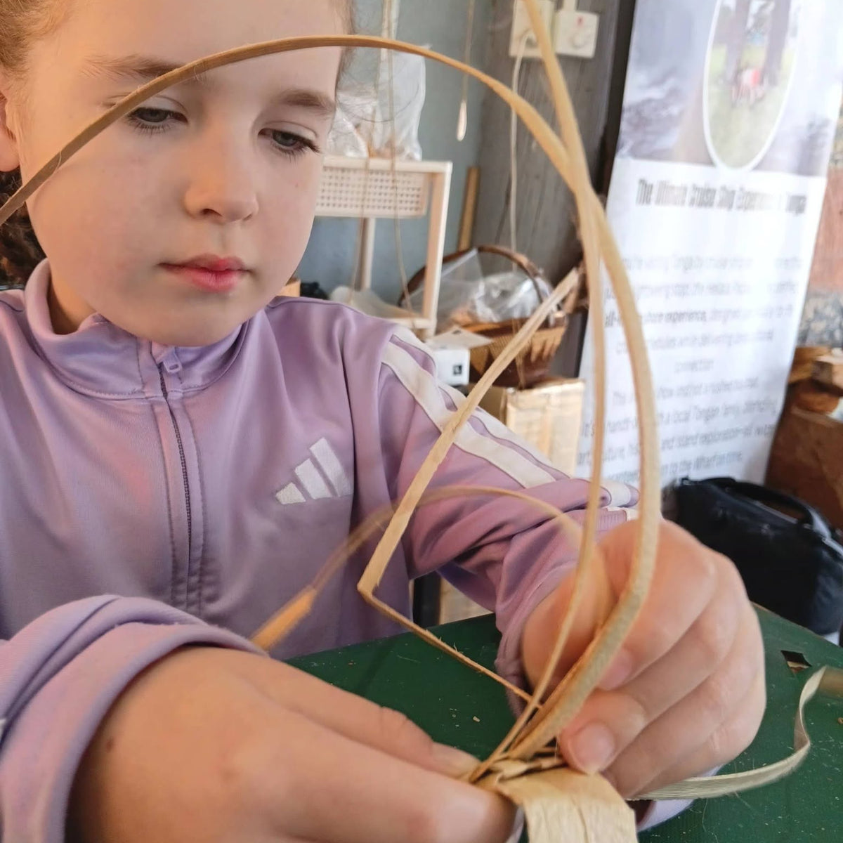 A young girl wearing a lavender Adidas zip-up jacket is focused on weaving a curved basket or tray from natural fiber strips over a green table.