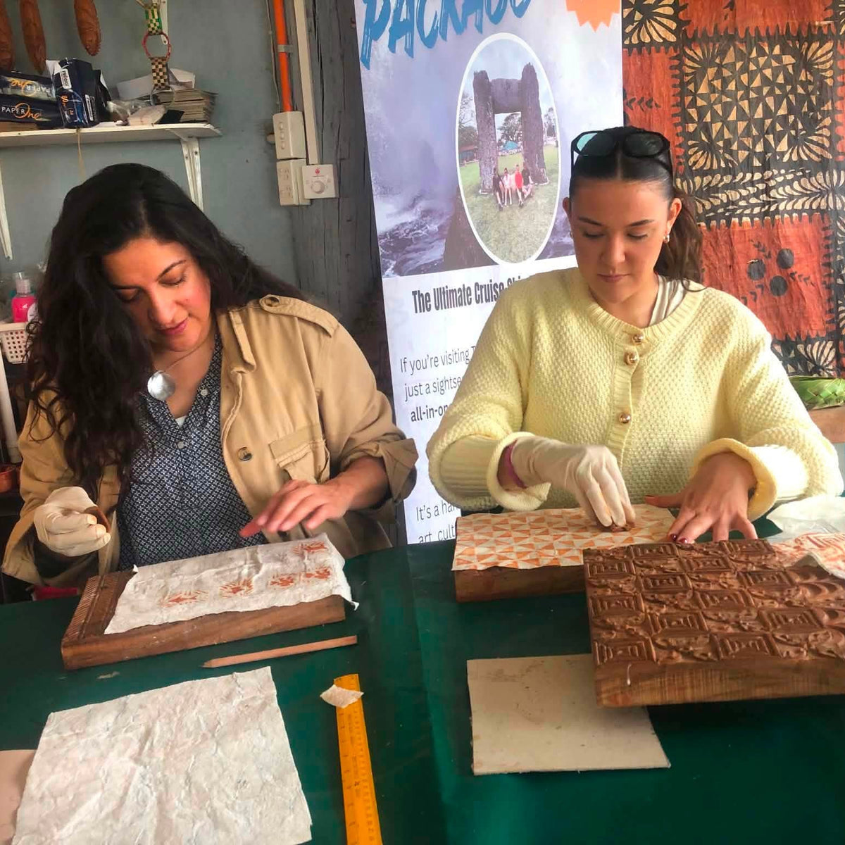 Two women seated at a table working with carved wooden blocks and paper, in a workshop-like setting with a patterned textile and printed poster in the background.