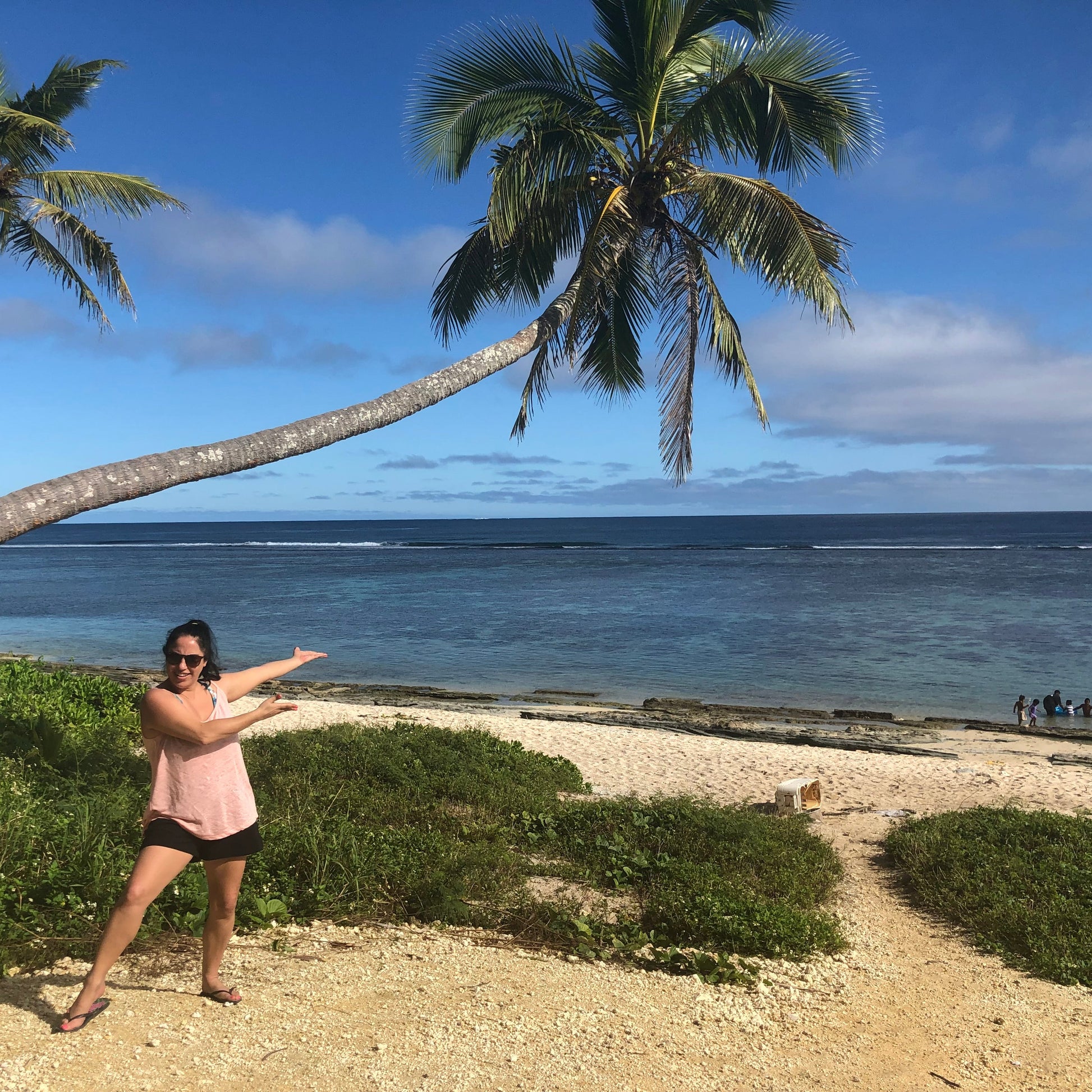 A woman posing on a tropical beach with palm trees, blue ocean, and a sandy shore in the background.