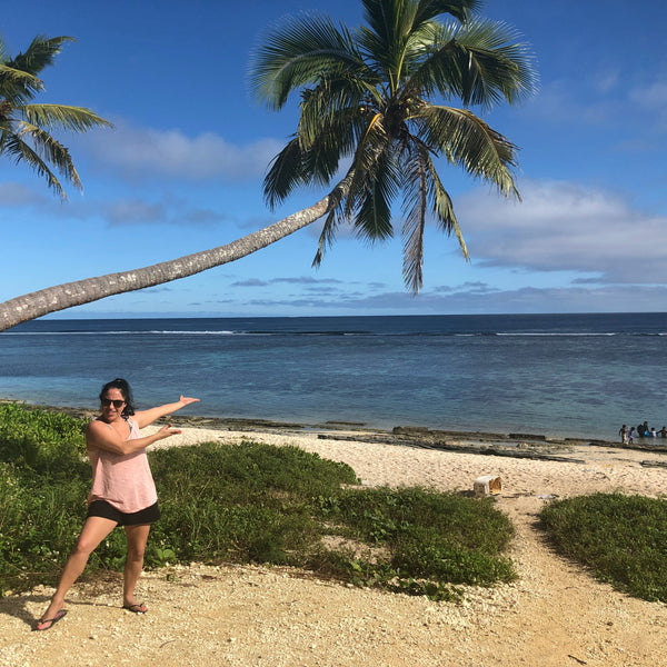 A woman posing on a tropical beach with palm trees, blue ocean, and a sandy shore in the background.