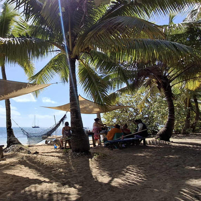 A sunny beach scene with palm trees, large tan beach umbrellas, sand chairs, picnic tables, and several people sitting and relaxing near the shoreline with boats in the water.