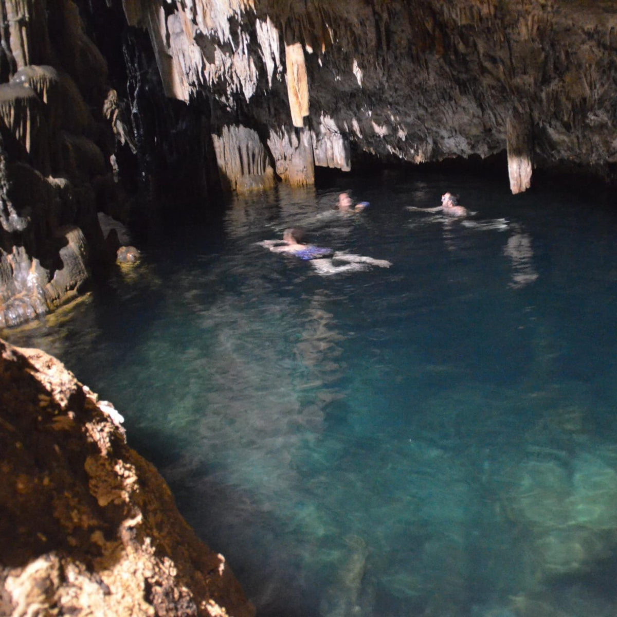 A scenic view inside a cave with clear turquoise water, stalactites, and stalagmites, with several people swimming in the pool.