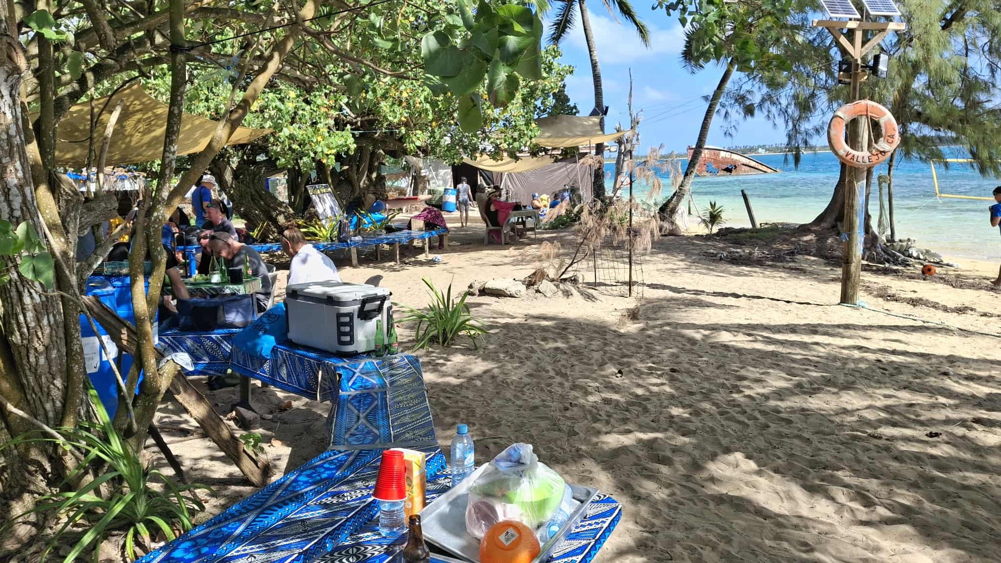 A beachside camping setup with blue folding tables, coolers, and gear under trees on sandy ground near the shoreline, with people visible in the background.
