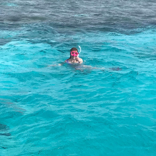 A person snorkeling in bright turquoise ocean water, wearing a pink snorkel mask and a white swim cap.