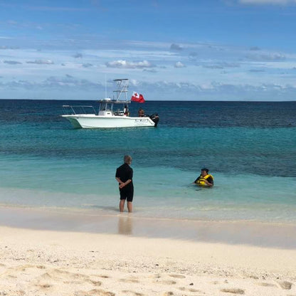 A beach scene with clear turquoise water, a white boat anchored offshore, and two people in the shallow water near the shore under a blue sky with scattered clouds.