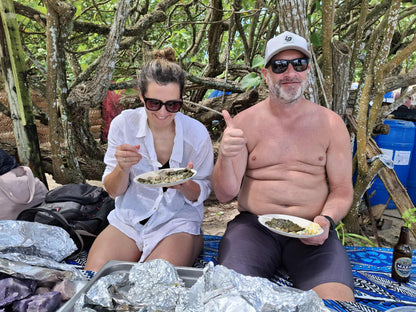 A man and a woman sitting outdoors on a blanket under trees, eating from paper plates. The man is shirtless and wearing a white cap and sunglasses, giving a thumbs-up.