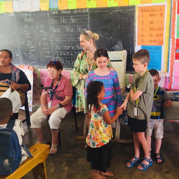A group of people, including children and adults, gathered inside a classroom with colorful educational posters on the wall and a chalkboard in the background.