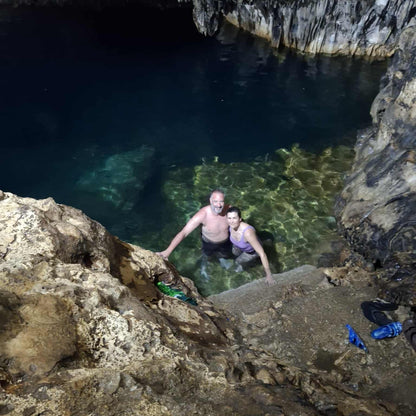 A couple standing in clear, shallow natural spring water inside a rocky cave, with dark blue water and cave walls surrounding them.