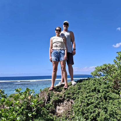 A couple posing on a grassy cliff overlooking the ocean under a clear blue sky. The woman wears a beige sleeveless top and denim shorts, and the man wears a white tank top, dark shorts, and a white cap.