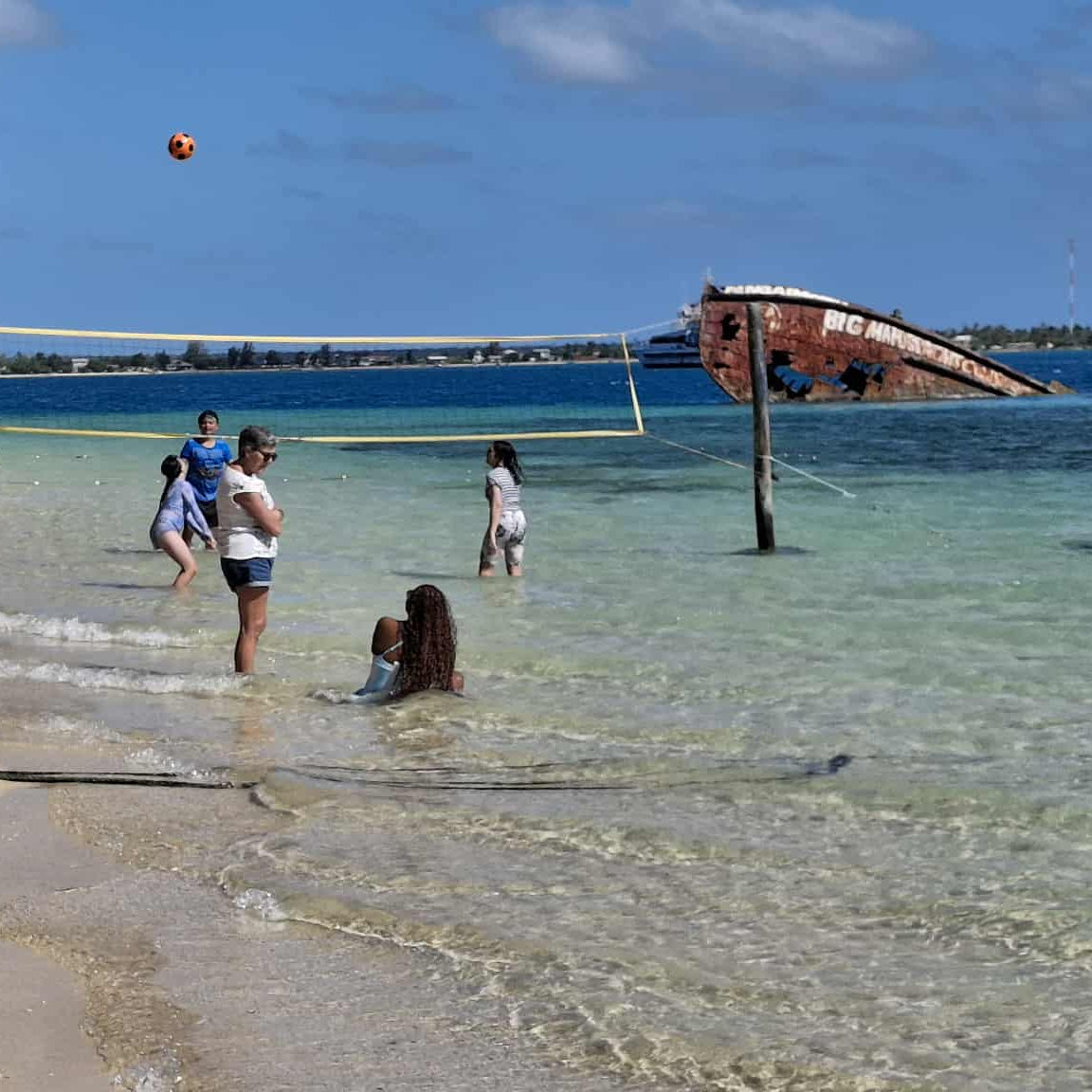 A beach scene with several people standing in shallow turquoise water near a volleyball net, with a shipwreck visible in the background under a blue sky.