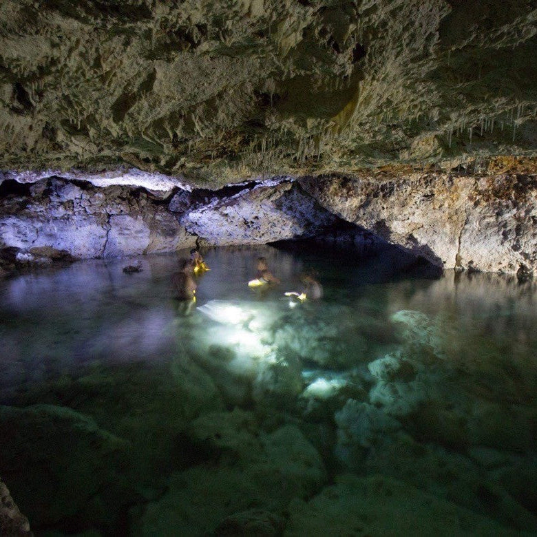 A cave interior with a clear pool of water reflecting artificial lighting, surrounded by rocky cave walls and ceiling.