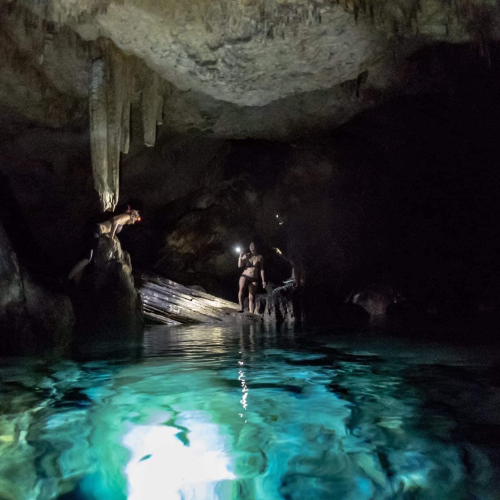 A person in a bikini stands in a dark cave holding a phone flashlight above glowing turquoise water, with stalactites and rock formations visible in the cave ceiling and walls.