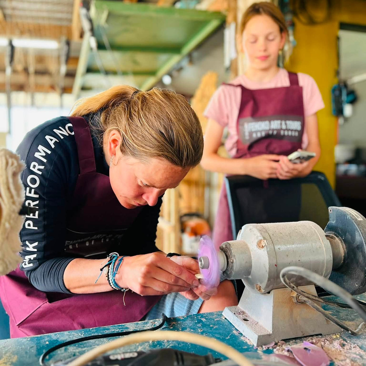 A woman in a maroon apron uses a rotary wheel grinder on a small purple object at a workbench, with another woman standing in the background inside a workshop.