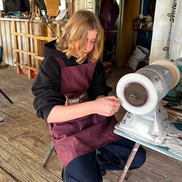 A person wearing a dark hoodie and a burgundy apron sits in a workshop, operating a bench grinder. The background shows wooden floors and shelves with various tools and materials.