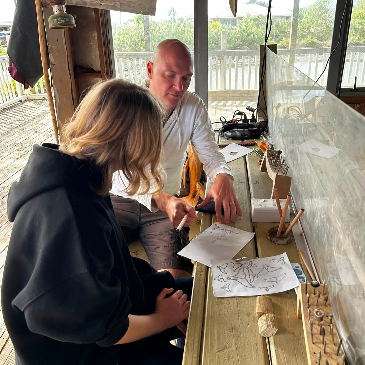 A man and a woman are seated at a wooden workbench in a rustic workshop, looking at and pointing to sketch papers. The man wears a white shirt and the woman wears a black hoodie.