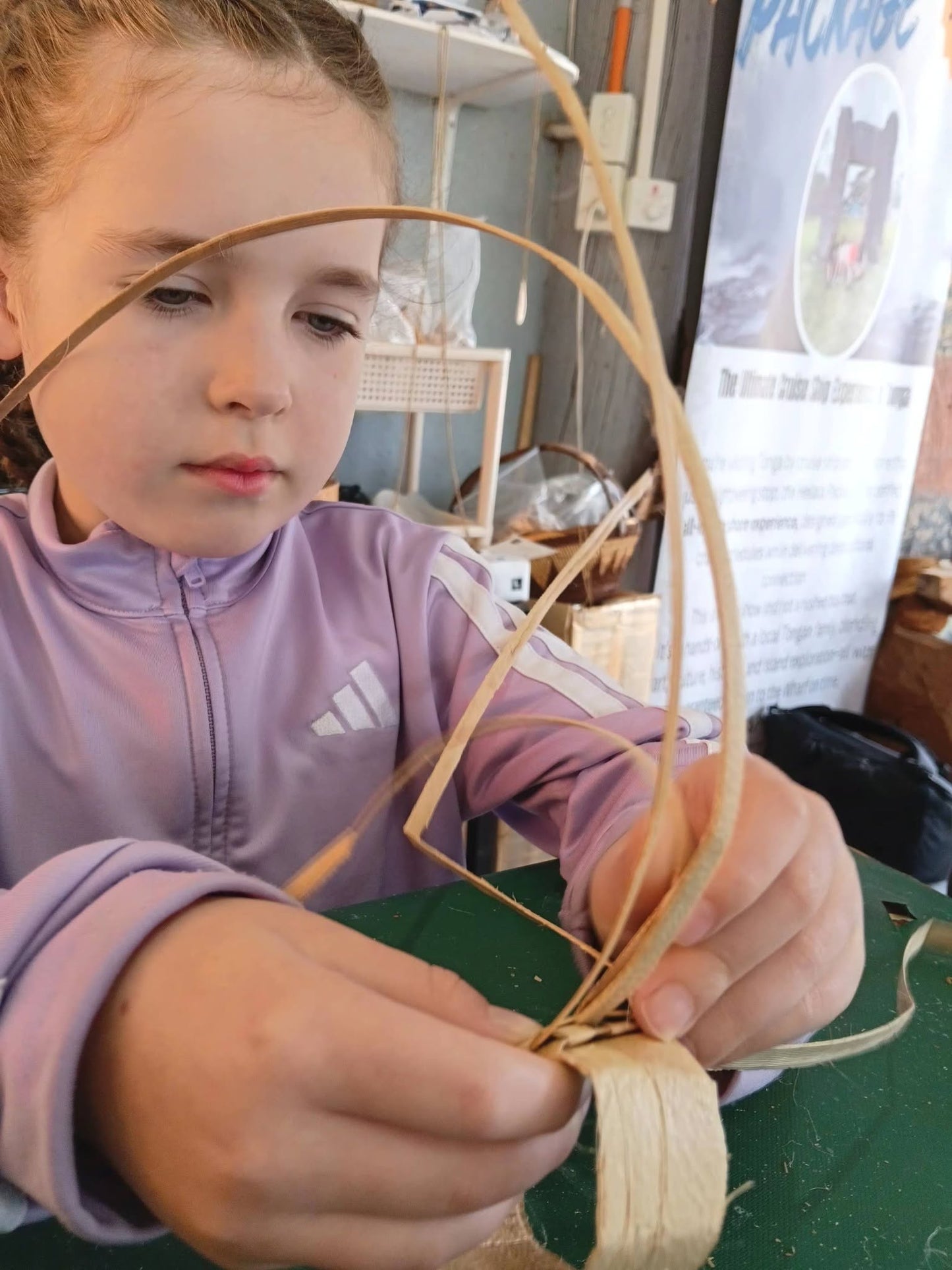 A young girl wearing a lavender Adidas zip-up jacket is focused on weaving a curved basket or tray from natural fiber strips over a green table.