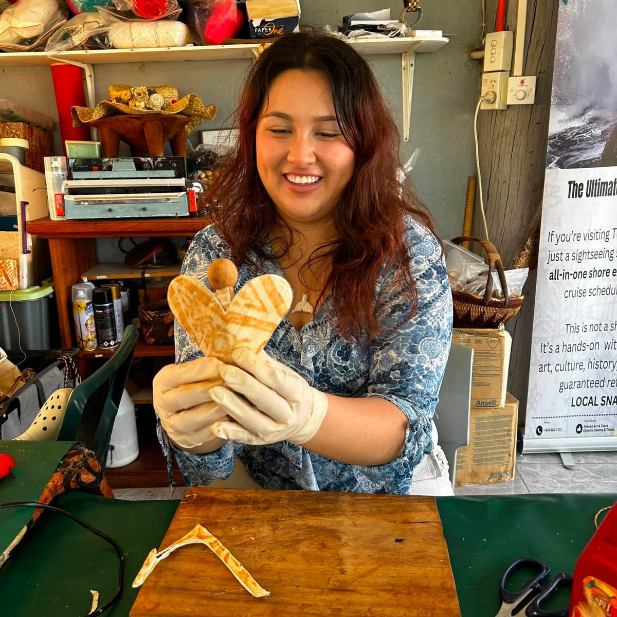 A smiling woman wearing gloves holds a handcrafted wooden heart-shaped ornament in a workshop filled with shelves, tools, and crafting supplies.