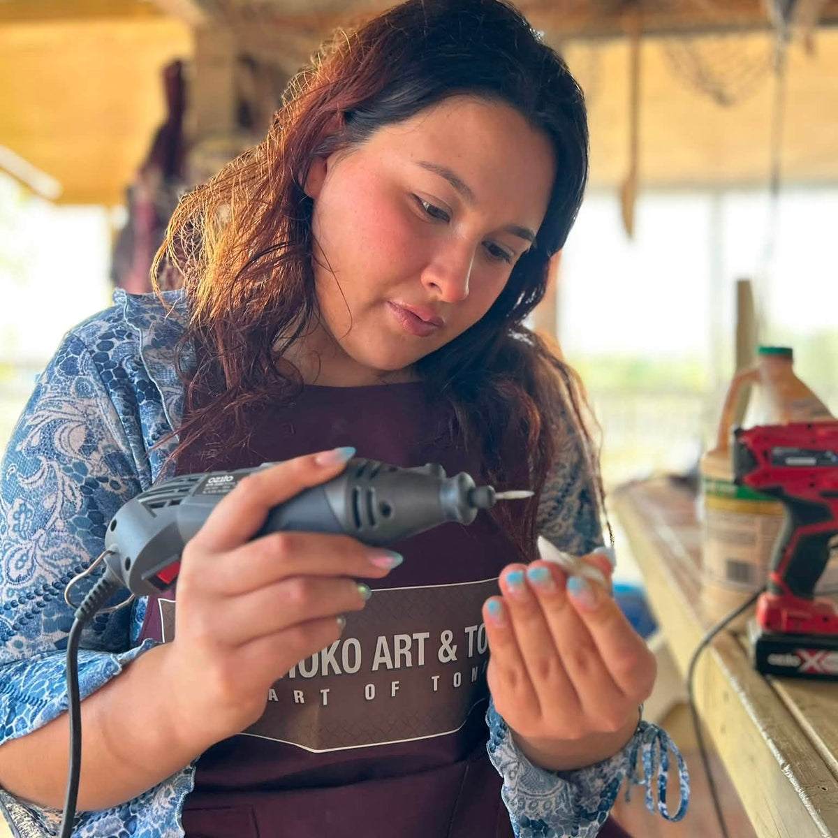 A woman wearing a maroon apron with 'KUKO ART & TOY' printed on it, using a rotary tool to work on a small object in a workshop setting.