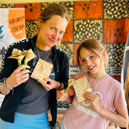 A woman and a young girl smiling while holding handmade paper goods, standing in front of a colorful patterned textile wall. The woman wears a black zip jacket over a gray shirt and blue skirt, and the girl wears a pink Hummel t-shirt and white shorts.