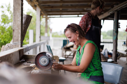 A woman in a green patterned sleeveless top smiles while sharpening a circular saw on a wooden porch. Another person is visible in the background under a covered deck.
