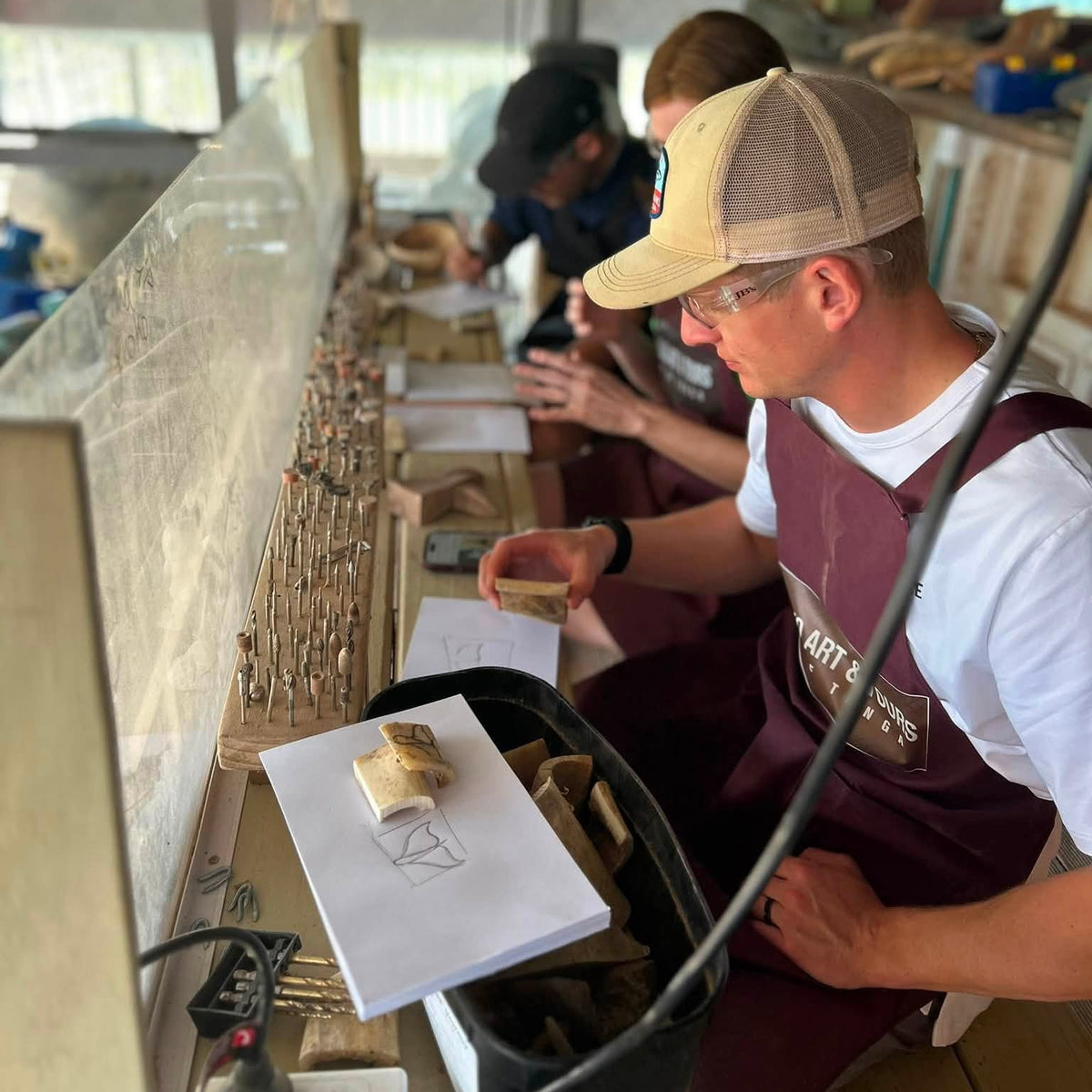 A man wearing a tan and brown trucker hat, white t-shirt, and maroon apron works at a wooden craft table inside a shop. Another person is visible in the background, and the table holds wood pieces, papers, and tools.