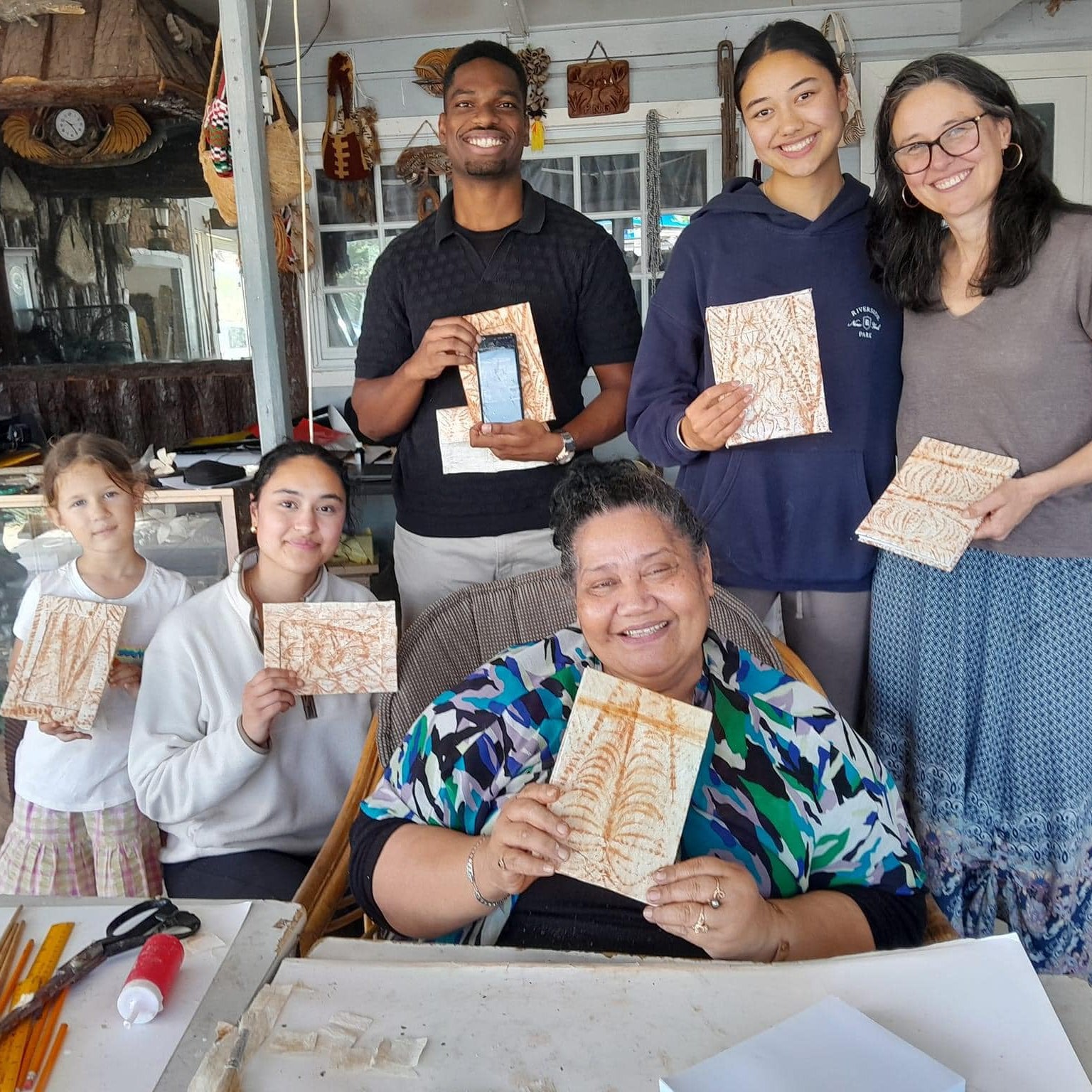 Six people smiling and holding handmade wooden art plaques inside what appears to be a craft studio or gallery.