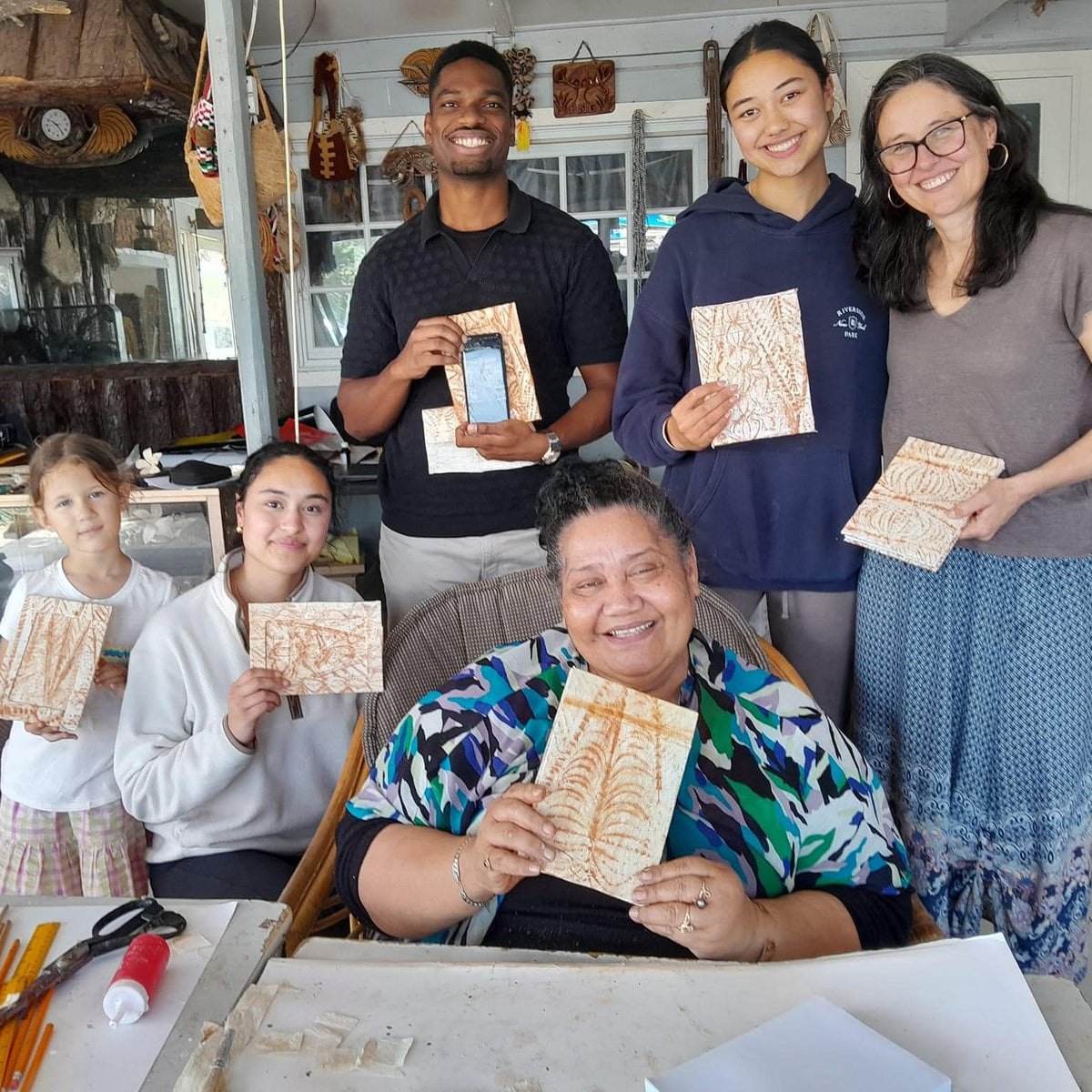 Six people smiling and holding handmade wooden art plaques inside what appears to be a craft studio or gallery.