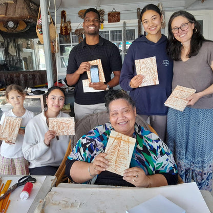 Six people smiling and holding handmade wooden art plaques inside what appears to be a craft studio or gallery.