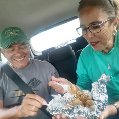 A man and a woman sitting inside a car, sharing a food item wrapped in aluminum foil. The man wears a green cap and gray t-shirt, and the woman wears a teal long-sleeve shirt and glasses.
