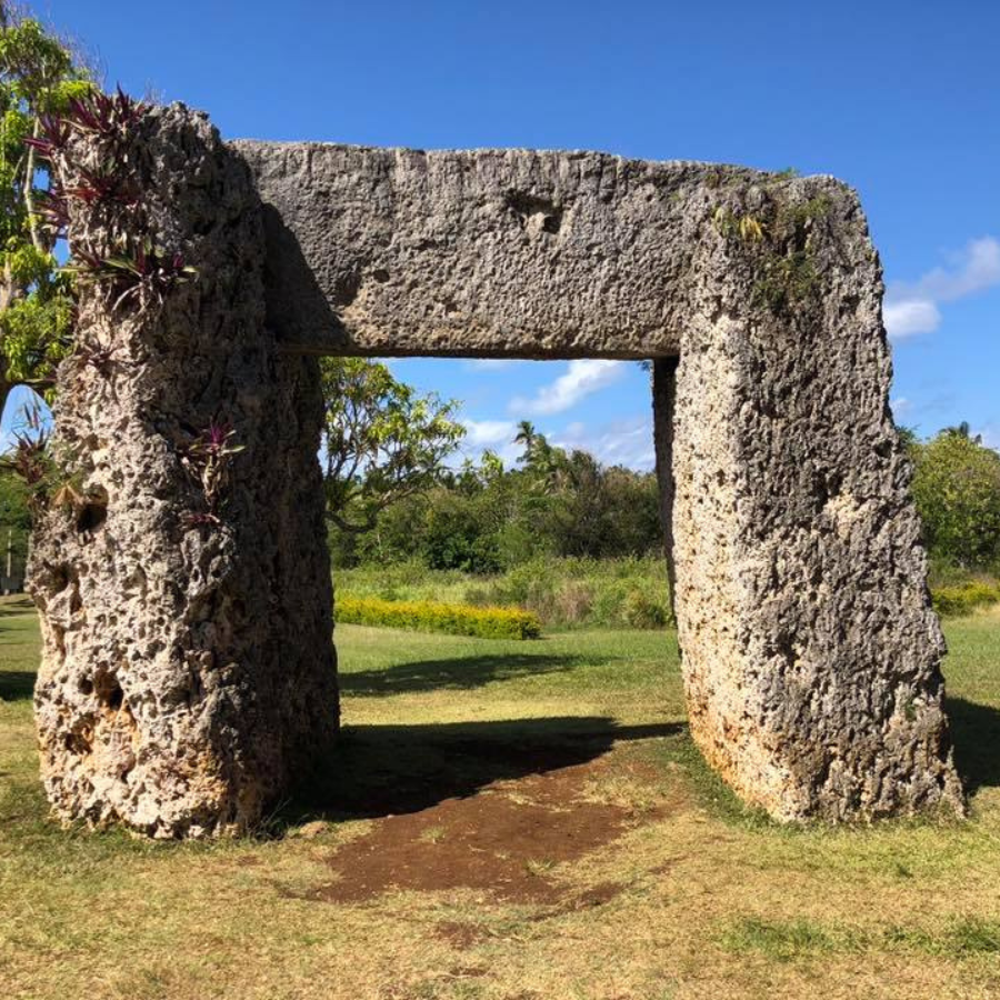 A large natural stone archway or trilithum structure standing on grass in an outdoor park, with trees, shrubs, and blue sky in the background.
