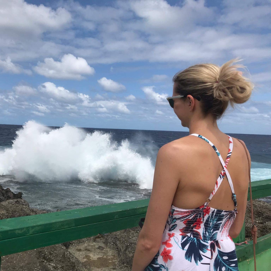 A woman in a tropical floral print halter dress stands on a rocky seaside overlook, looking out at a large crashing ocean wave under a cloudy blue sky.