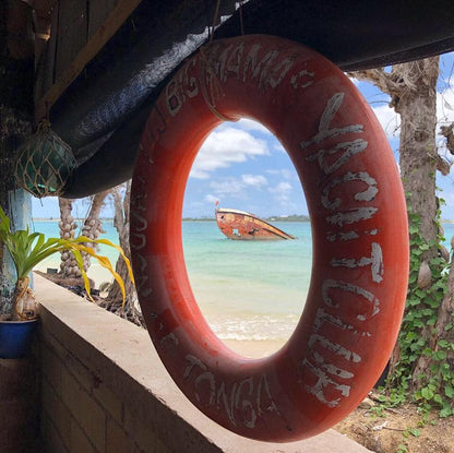 A red lifebuoy with the text 'Night Club Tonga' hung on a wall, with a body of water and a boat in the background.