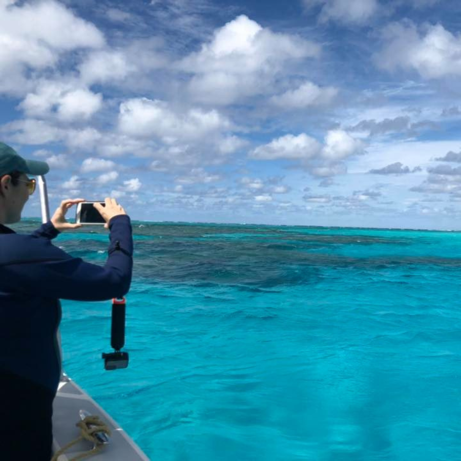 A person in a dark wetsuit and cap takes a photo with a smartphone from a boat, overlooking vibrant turquoise ocean water under a blue sky with scattered white clouds. A small tropical island is visible in the distance.