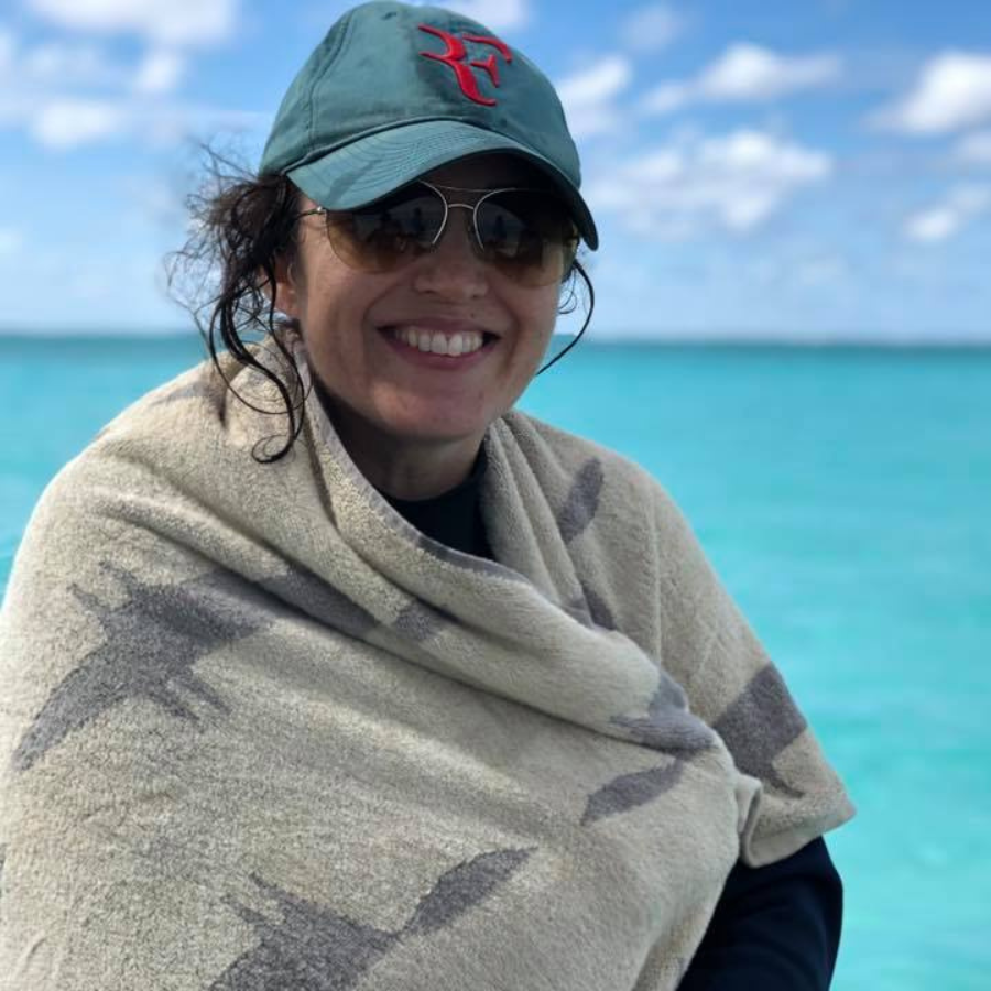 A smiling woman outdoors by the turquoise ocean, wearing a green baseball cap with a red embroidered logo, sunglasses, and a beige patterned wrap or blanket draped around her shoulders.