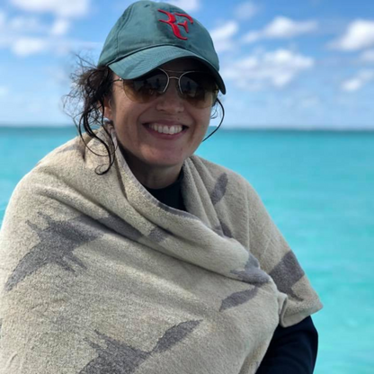 A smiling woman outdoors by the turquoise ocean, wearing a green baseball cap with a red embroidered logo, sunglasses, and a beige patterned wrap or blanket draped around her shoulders.