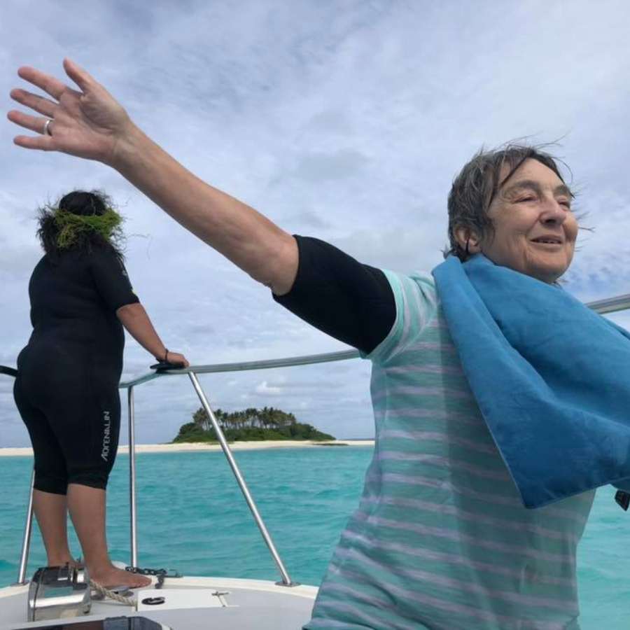 A woman on a boat with her arm raised in the air, wearing a light blue and white striped shirt and a bright blue towel draped over her shoulder. Another person in a black swimsuit stands in the background on the boat against a tropical ocean and cloudy sky.