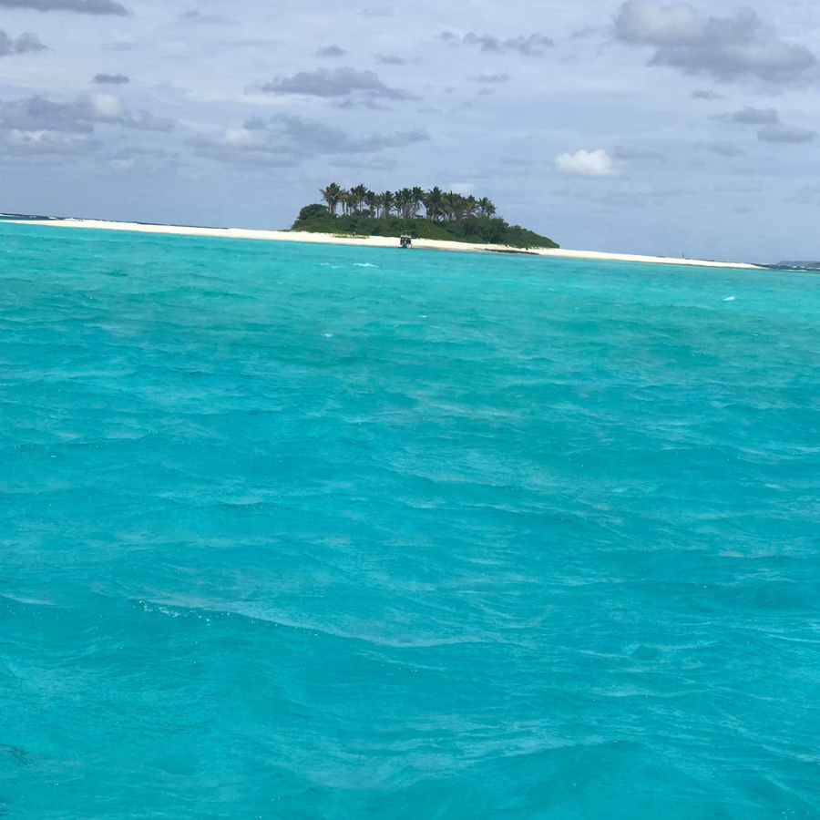 A scenic view of a tropical island with a long white sandbar and palm trees, surrounded by bright turquoise ocean water under a cloudy sky.