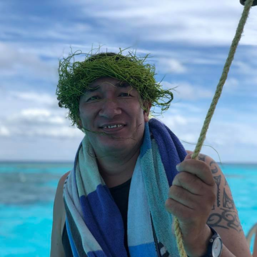 A man smiling on a boat with turquoise ocean water and a cloudy blue sky in the background. He is wearing a green seaweed head wrap and a blue, white, and light blue striped towel around his neck.