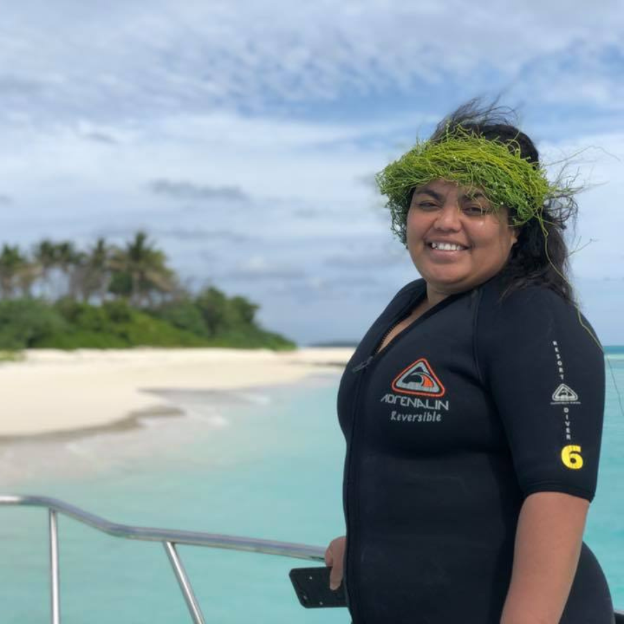 A woman wearing a black wetsuit with 'AIDAUN Everest' printed on the chest stands on a boat deck with a tropical beach and palm trees in the background.
