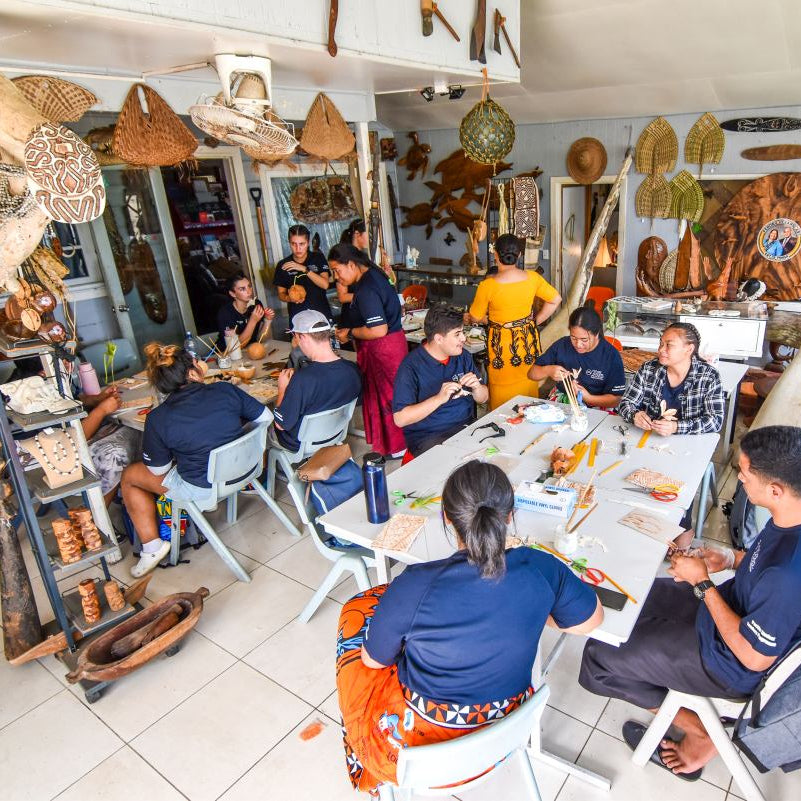 A group of people seated around a large white table inside a shop filled with handcrafted wooden and natural items, including baskets, carved decor, and hanging tools.