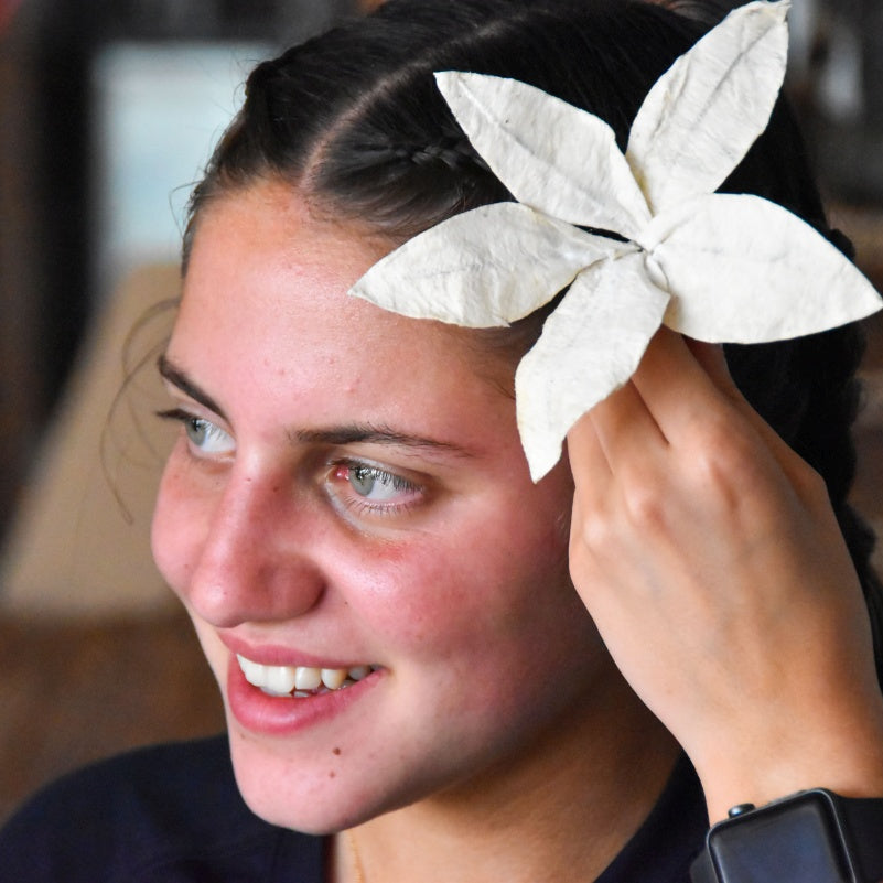 A close-up lifestyle shot of a woman with dark hair wearing a white paper flower hair accessory, holding it in place with her hand.