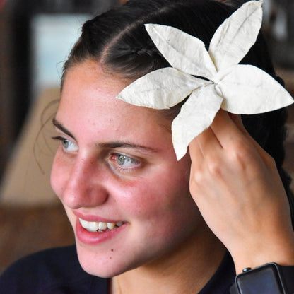 A close-up lifestyle shot of a woman with dark hair wearing a white paper flower hair accessory, holding it in place with her hand.