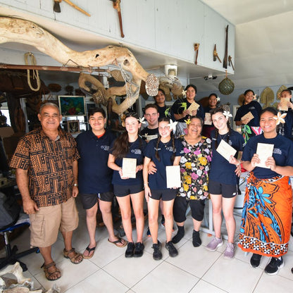 A group of people standing together inside a shop or workshop, holding small white certificates. Several individuals are wearing matching dark navy blue polo shirts with white logos, while others wear casual clothing.