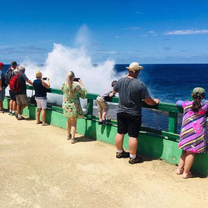 A group of people standing on a pier or deck overlooking the ocean, with a large sea spray plume erupting in the blue sky. The people are dressed in casual summer clothing, and the scene is set outdoors with no visible product being sold.