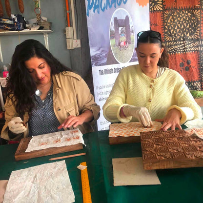 Two women seated at a table working with carved wooden blocks and paper, in a workshop-like setting with a patterned textile and printed poster in the background.