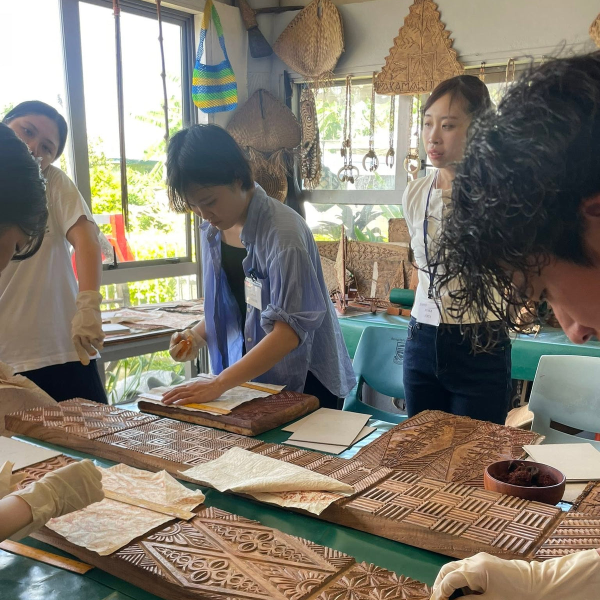 Several people gathered around a table in a workshop, wearing gloves and working with carved wooden blocks and printed cloth sheets. The scene shows a real indoor environment with windows, hanging woven items, and tools, so this is Lifestyle rather than Product/Packaging.