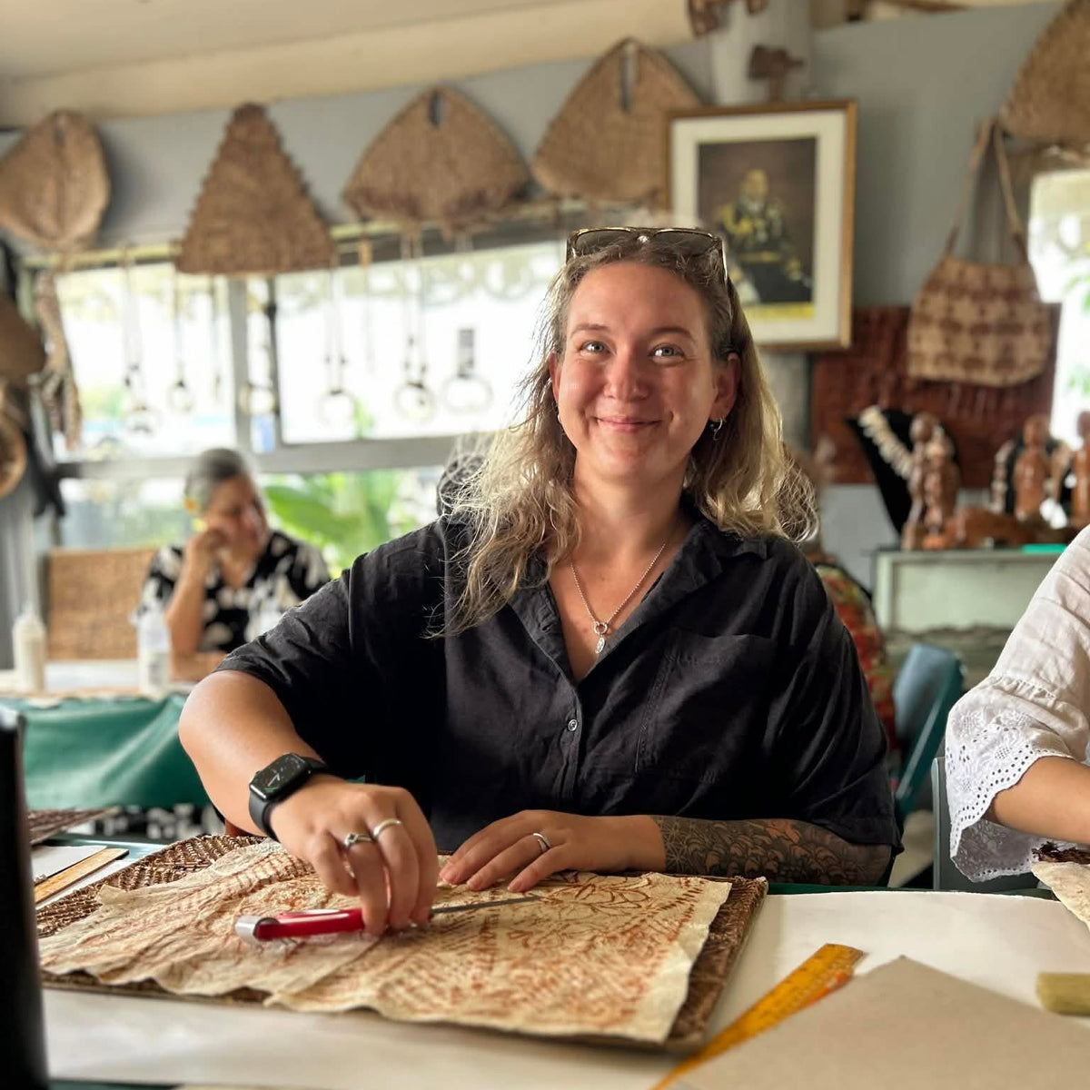 A woman with blonde hair sitting at a table in a shop, smiling and looking at a piece of patterned fabric or paper. She is wearing a black button-up shirt.