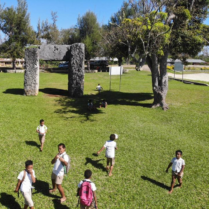 A grassy outdoor site with large stone monoliths in the background and several children walking across the lawn under a blue sky.