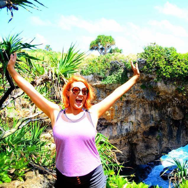 A woman with red hair and sunglasses stands outdoors with her arms raised, smiling in front of a rocky coastal cave with blue water below and lush green tropical plants.