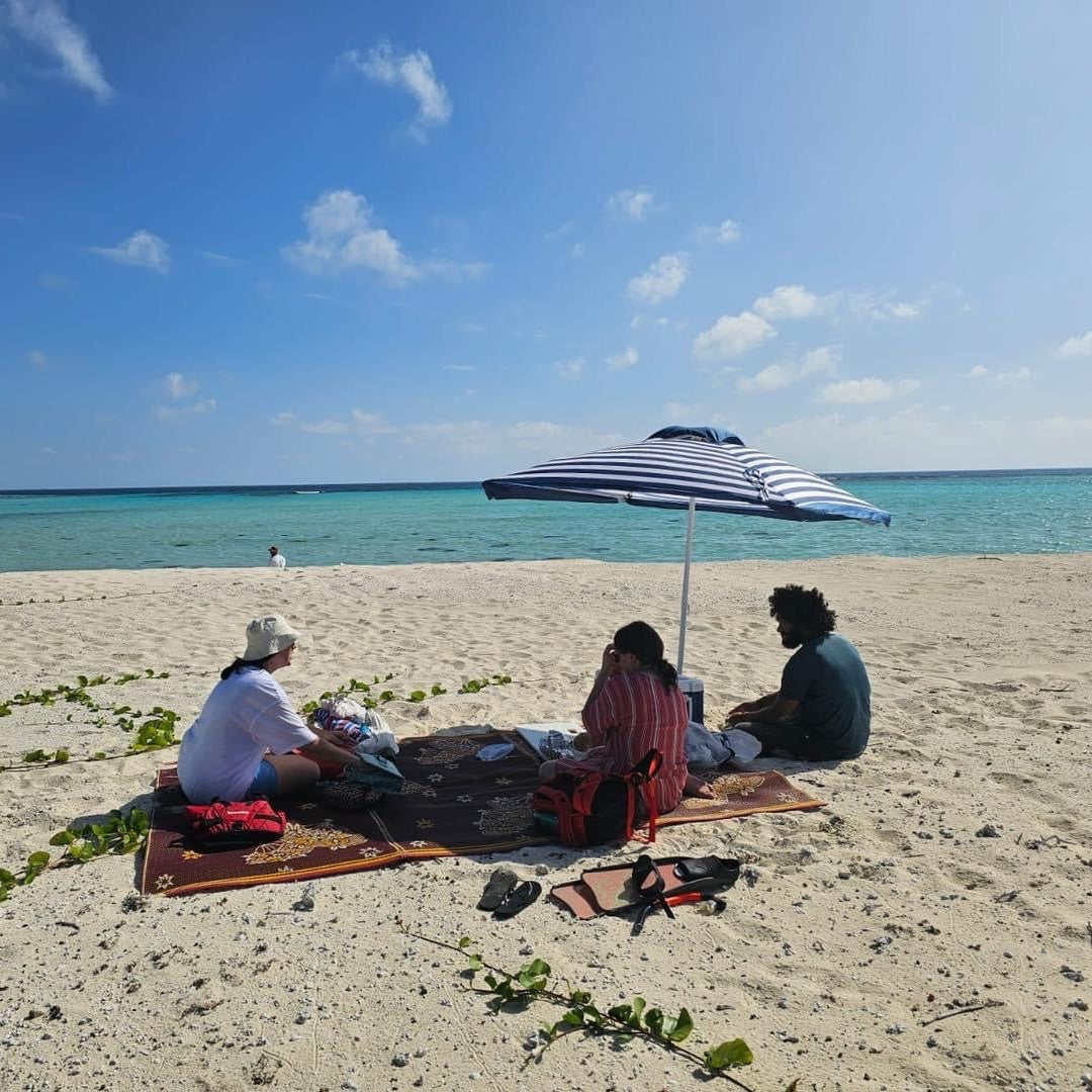 Three people sitting on beach towels under a striped blue and white umbrella on a sandy beach, with turquoise ocean water and a blue sky in the background.